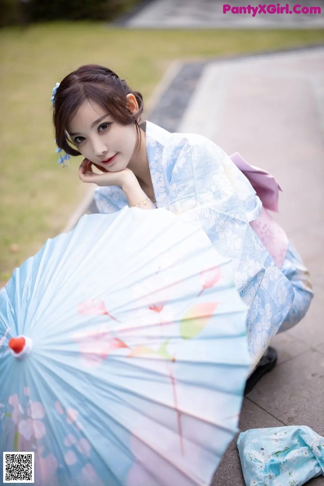 A woman in a blue kimono sitting on the ground with an umbrella.